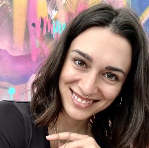 Cropped square photo of a headshot of artist Meaghan Kehoe, they are smiling at the camera. Meaghan has medium-length brown hair, light-medium skin and is wearing a black shirt and jewellery. They are in front of a colourful mural background.