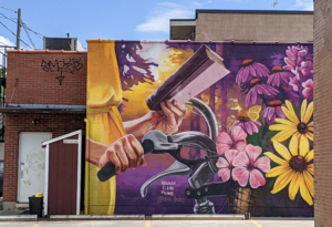 Horizontal photo of a mural by Meaghan Kehoe on an exterior building. It features a close crop of a person holding a book and holding onto the handles of a bicycle with a basketful of pink and purple flowers.