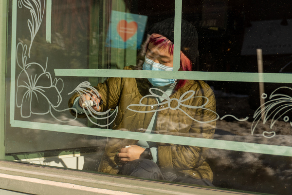 Photograph of an artist drawing a mural on a storefront window with a white paint marker. The mural features floral patterns drawn within a green tape border along the sides of the window.
