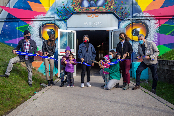 Photograph of a group of people standing in front of a large-scale mural painted across the entrance of a TTC station. The mural features imagery of bird eyes surround by brightly coloured rays surrounding it. 