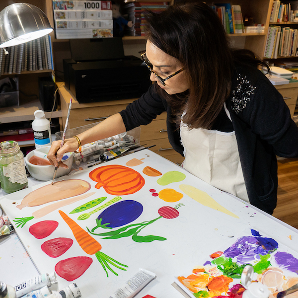 Photograph of artist Farida Zaman painting in her studio on a canvas, surrounded by paints and books. The painting features imagery of fruits and vegetables. 