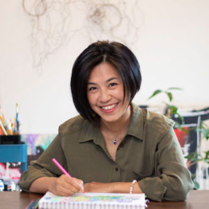 Jeni Chen smiling at the camera while seated at a table in an art studio. She is Asian with a black bob haircut and wearing an olive green collared shirt.