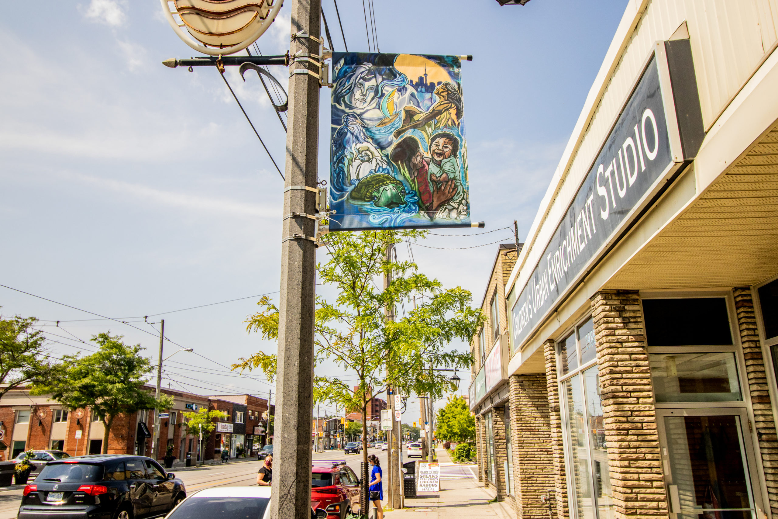 A street pole banner artwork by artist Danielle Hyde on a sunny day. The artwork is a digital piece with illustrations inspired by Toronto's waterfront with images of the cityscape, families and animals