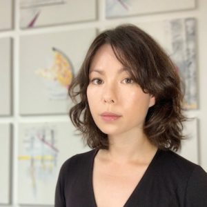 Meghan Cheng is looking at the camera with artwork hung behind her. She is of mixed-Asian race with short, wavy brown hair and wearing a black v-neck top.