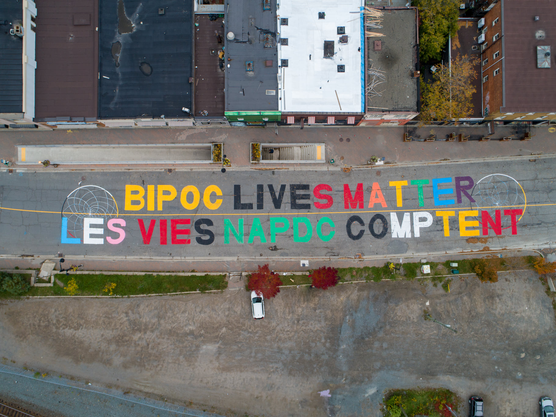 An aerial shot of a large ground mural spanning a road with colourful block letters "BIPOC LIVES MATTER" and underneath is the French version "LES VIES NAPDC COMPTENT"