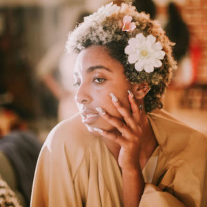 Ra'annaa Brown looking off to the side and posing with her face resting in her hands. Her short hair is adorned with flowers and she is wearing a brown, flowy top.