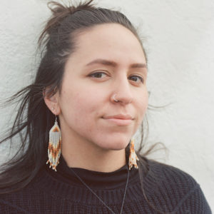 Stephanie Babij smiling at the camera. She is an Indigenous artist with brown hair tied back in a half up-do. She is wearing dangly beaded earrings and a dark sweater.