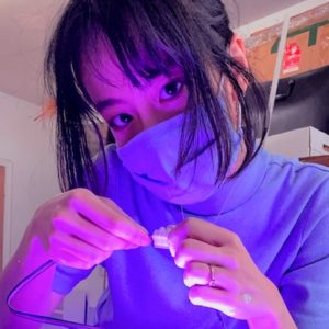 Tina Nguyen wearing a face mask and working on an art project. She is Asian with dark hair and bangs and in a room illuminated in purple lighting.