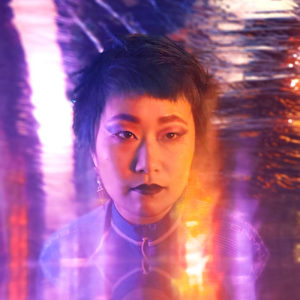 Artist Enna Kim of Korean-Canadian descent who has a pixie haircut and wearing dark lipstick and long earrings. Enna is photographed under dreamy purple and pink lighting.