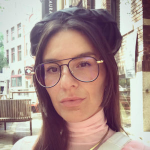 Niki Little gazing at the camera. She has long straight brown hair and is wearing a beret. She is also wearing aviator glasses with a pink turtleneck shirt.