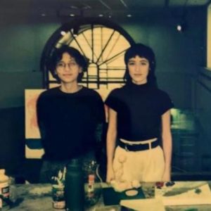 RAZA Collective with Laura and Valentina Caraballo standing side by side. Valentina has short wavy hair with glasses and wearing a black long sleeve. Laura has shoulder length hair with bangs and wearing a black short sleeve. They are both standing in front of a table with art materials