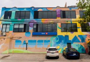 Photograph of the rear of a building overlooking a parking lot with cars parked. The building facade has a large-scale mural featuring imagery of shapes and stylised font painted in vibrant colours.