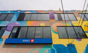 Close up photograph of the top of a building that features a large scale mural and a sign that reads "BMO". The mural is features imagery of brightly coloured shapes and patterns.