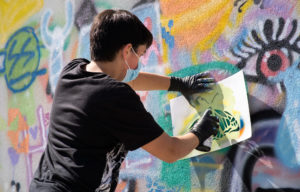 Photograph of a participant from the workshop spray painting on a stencil, adding to the art already painted on the wall.