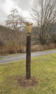 Vertical photo of a bronze sculpture that is sculpted into a cedar cone shape. There is grass, grey skies, a road and trees surrounding it.