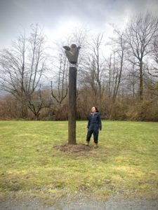 Vertical photo of a bronze sculpture that is sculpted into a cedar cone shape, held up by a log of wood, and placed in the center, artist Laara Cerman stands beside it smiling. There is grass, grey skies and trees surrounding it.