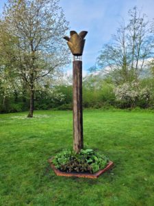 Vertical photo of a bronze sculpture that is sculpted into a cedar cone shape, held up by a log of wood, and placed in the center of a small garden. There is grass, shrubery, blue skies and trees surrounding it.