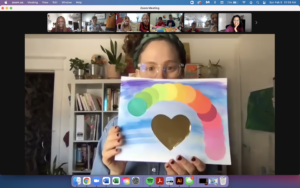 Photograph of a virtual art class. The instructor, Bareket Kezwer, is on the largest frame, while the participants are in smaller frames at the top of the screen. Bareket is holding up an artwork featuring imagery of a gold heart surrounded by an arc of colourful circles set against a blue background.
