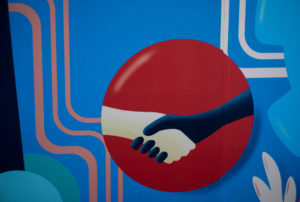 Close-up photograph of an interior mural focusing on two clasped hands enclosed within a red circle set against a bright blue background with line patterns.
