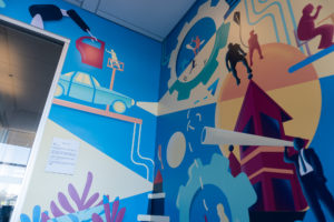Close-up photograph of an interior mural, focusing on the upper half. The mural features detailed imagery of figures, buildings, cars and other patterns set against a bright blue background.