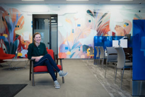 Artist Mary Haasdyk poses in front of her interior mural with bright teal, orange, and red tones in a BMO office.