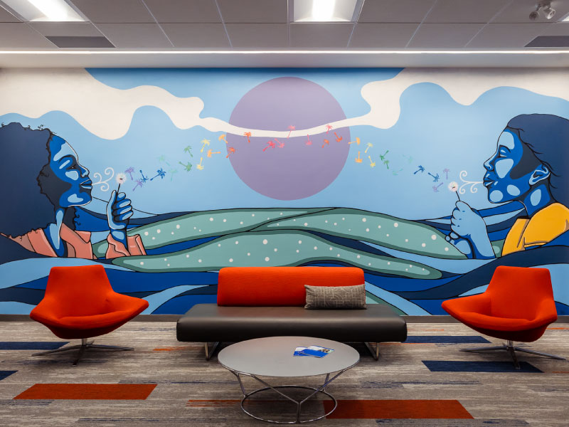 An interior vinyl mural by Mo Thunder in a bank branch as part of the BMO National Mural Series by STEPS Public Art. It depicts a scene of two people blowing dandelions across a sunset landscape.