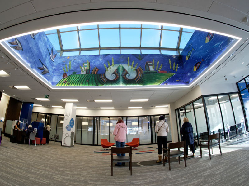 An interior digital vinyl mural wrapping around a skylight of a bank by artist Bruno Hernani as part of the BMO National Mural Series by STEPS Public Art. The artwork depicts Indigenous imagery, flying geese, and fish under water