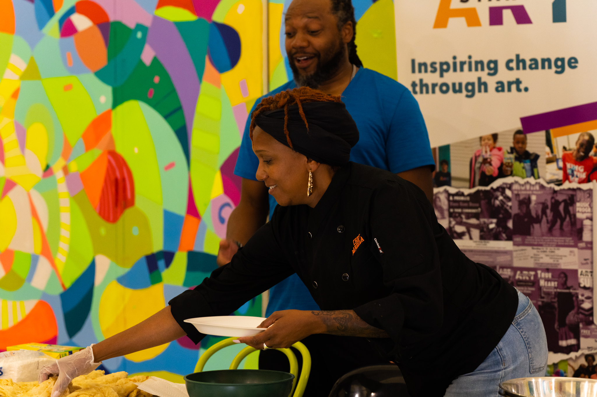 A Black chef reaching over a table to put food on a plate. There is a colourful mural in the background and a banner that says "Inspiring change through art"
