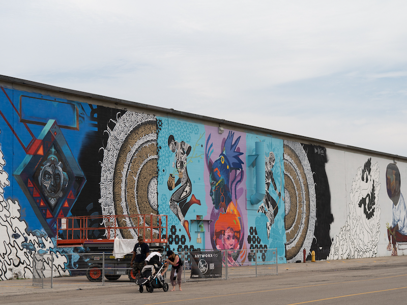 A portion of a 360 foot long mural production featuring ten different wall sections of colourful artwork by different artists. A section is fenced off with an active production and a lift. Two people are walking by with a stroller. 