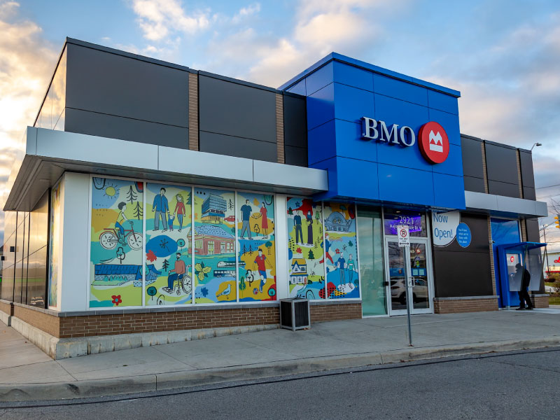 The entrance of a bank with a vinyl mural installation on the windows that showcase artwork of the local community doing activities. This is part of the BMO National Mural Series by STEPS Public Art