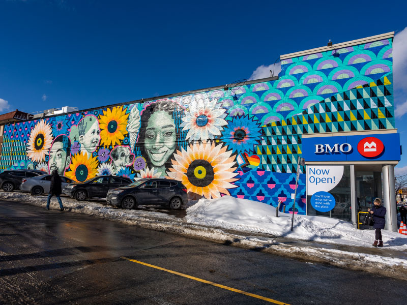 A large painted exterior mural on the side of a bank by artist Gosia Komorski as part of the BMO National Mural Series by STEPS Public Art. Sunflowers are surrounded by faces of community members.