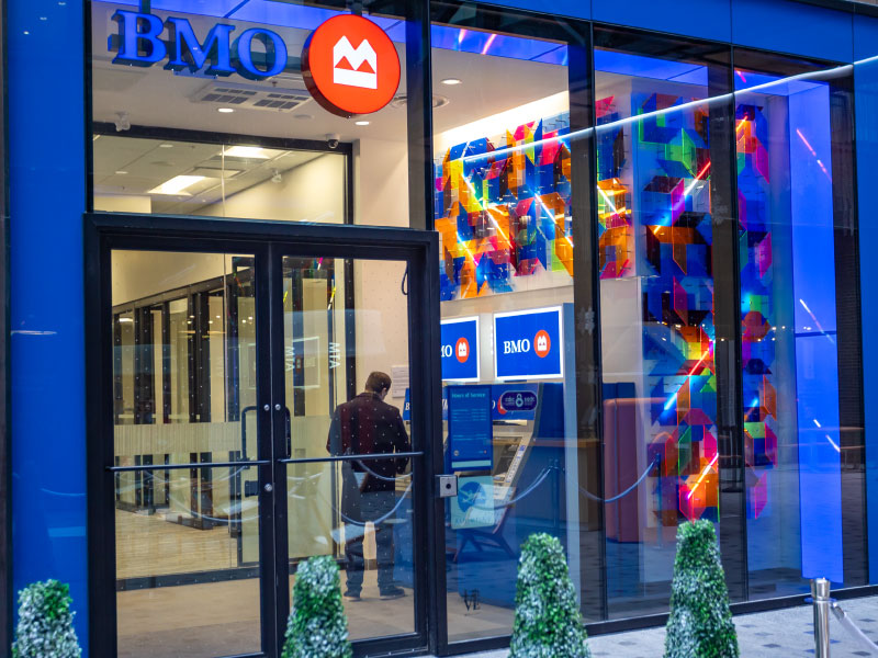 The exterior of a bank and a person using the ATM machine. There is a bright 3D geometric sculpture along the interior wall as part of the BMO National Mural Series by STEPS Public Art