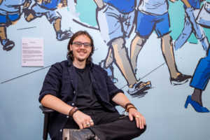 Photograph of artist Matt Salisbury sitting in front of his interior mural and curatorial panel that describes the inspiration for the artwork.