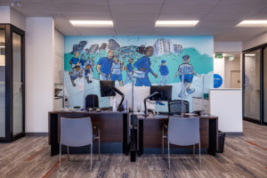 Photograph of an interior mural featuring imagery of figures walking on a path in an urban area painted in shades of blue and green installed in a BMO office.