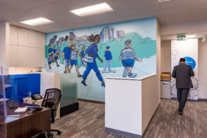 Photograph of a person walking past an interior mural featuring imagery of figures walking on a path in an urban area painted in shades of blue and green installed in a BMO office.