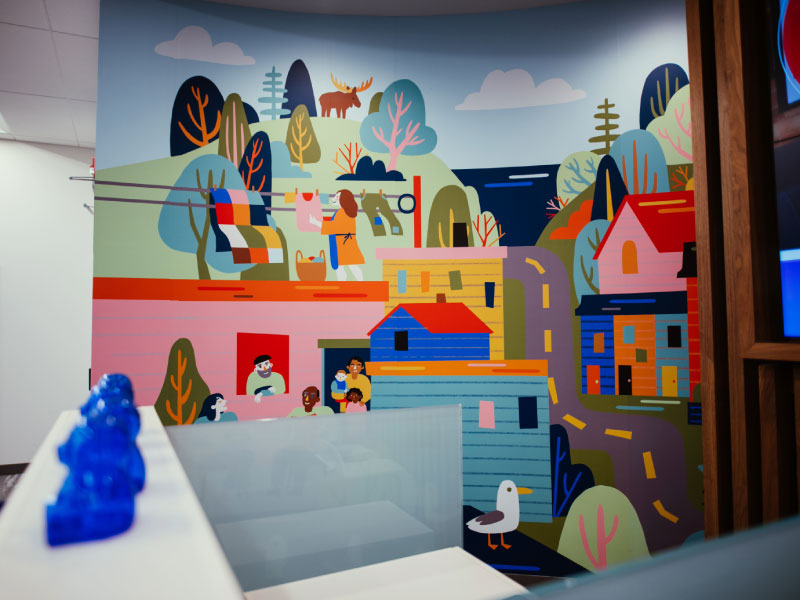 An interior digital vinyl mural by artist Molly Margaret as part of the BMO National Mural Series by STEPS Public Art. In a flat icon style with bold colours, it is a scene of a community with water and wildlife in the distance.