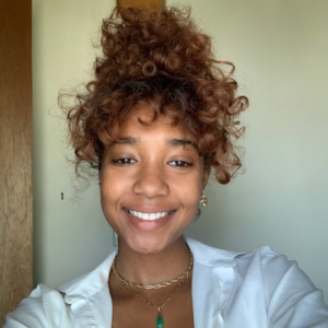 Renee Jagdeo smiling at the camera. She has light brown skin with curly brown hair tied on top of her head. SHe is wearing a white collared shirt with a gold necklace with a jade pendant.