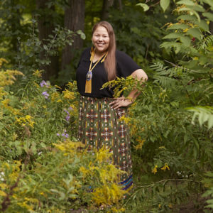 Lindsey Lickers, an Indigenous artist, standing in a park with trees and bushes surrounding her. She is smiling and posing at the camera. She has long brown hair and is wearing a black top, a long skirt, and a necklace.