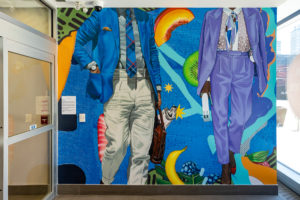 Photograph of a finely detailed interior mural featuring imagery of two figures in suits surrounded by patterns of fruit and other elements set against a bright blue background.