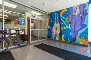 Photograph of a finely detailed interior mural featuring imagery of two figures in suits surrounded by patterns of fruit and other elements set against a bright blue background. Also seen is the entrance to the bank, with a bicycle leaning to the side.