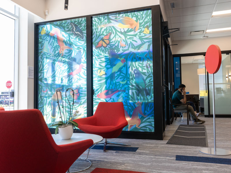 An interior translucent vinyl mural inside of a bank by artist Stephanie Scott as part of the BMO National Mural Series by STEPS Public Art. It depicts swirls of branches, birds, butterflies, and squirrels