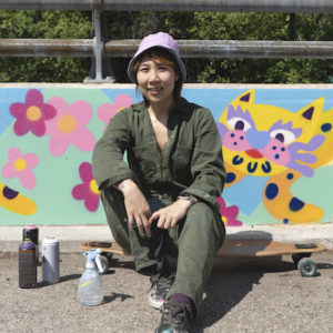 Artist Jieun June Kim sitting on a longboard in front of a guardrail mural of a tiger and flowers on a sunny day in Cooksville, Mississauga