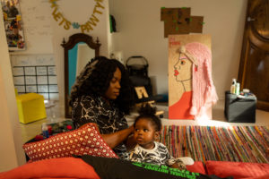 Photo of a Black woman wearing a printed top doing a young child’s hair, they are sitting on a red couch with pillows. The space they are seated in has various decorations such as a mirror, rug and banner. Also, it shows the artwork created for The Mane Event project in the back with a side portrait and pink braided hair.