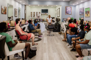 Photo of hair stylist TK at the front of a room, behind a chair, speaking to a group of hair workshop participants in a hair salon. The participants sit along both walls with various styling equipment, stations and decor on the walls.