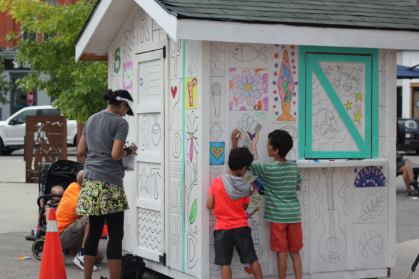 A family interacting with a wooden house that has colouring sheets hung up as part of a community engagement in Stratford, Ontario