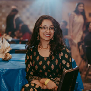 Headshot of artist Shivani Joshi smiling and wearing traditional Indian clothes, sitting in front of a table covered with a blue tablecloth.