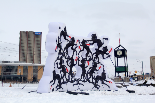 Large white inflatable public art installation by Paul Robles with dark silhouettes of playful monkeys with red hands. The installation is surrounded by snow and a clock tower in the City of Waterloo