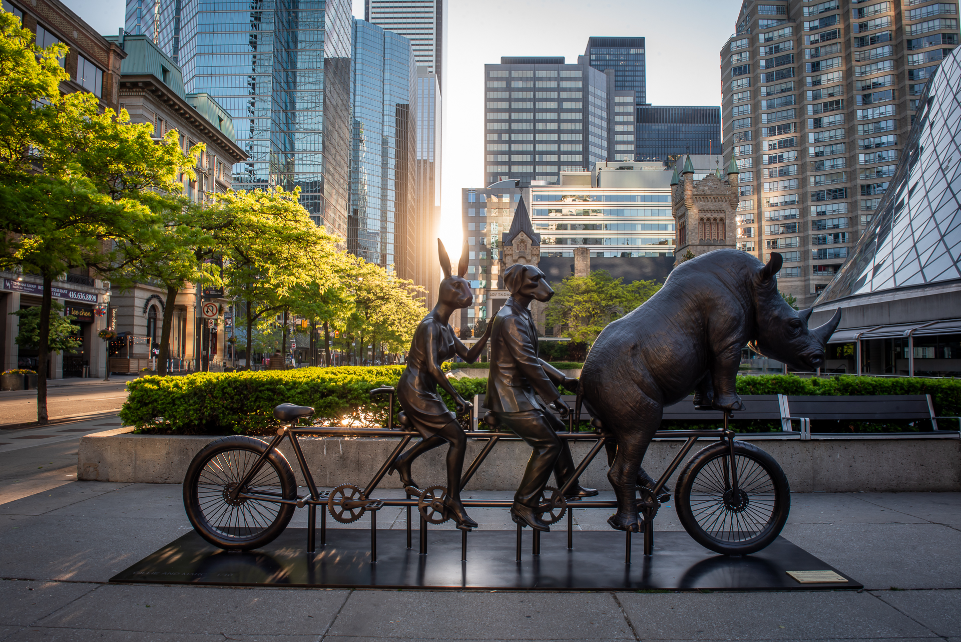 A sculptural installation of Rabbitwoman, Dogman, and a northern white rhinoceros on a bicycle in David Pecaut Square