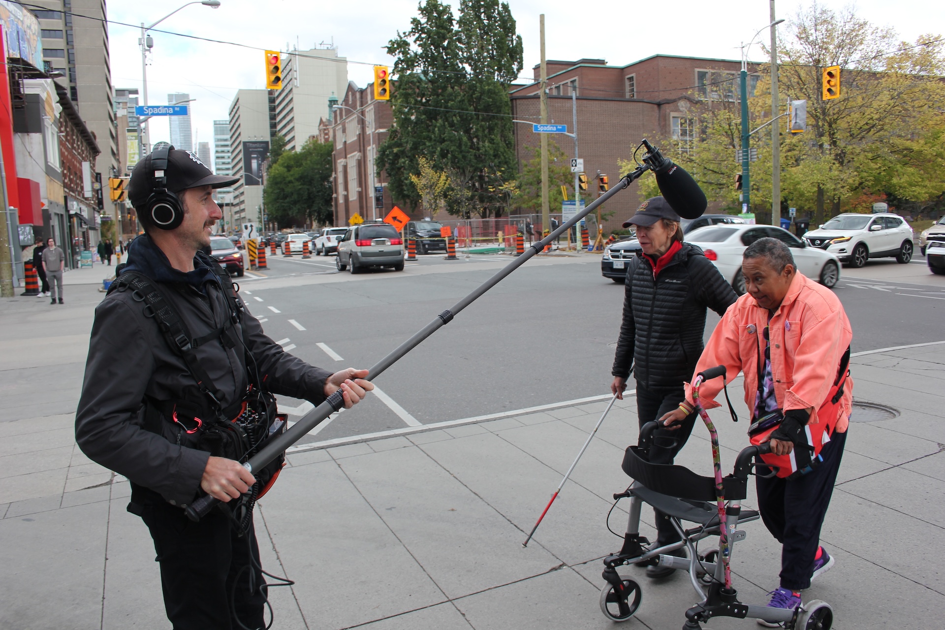 A senior using a walker is crossing the street with Blind artist and playwright Alex Bulmer. A person is recording the interaction with a boom mic. 