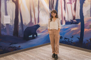 A mural of a black bear in a sunlit forest for the BMO National Mural series with artist Marcelline Siu standing in front of it